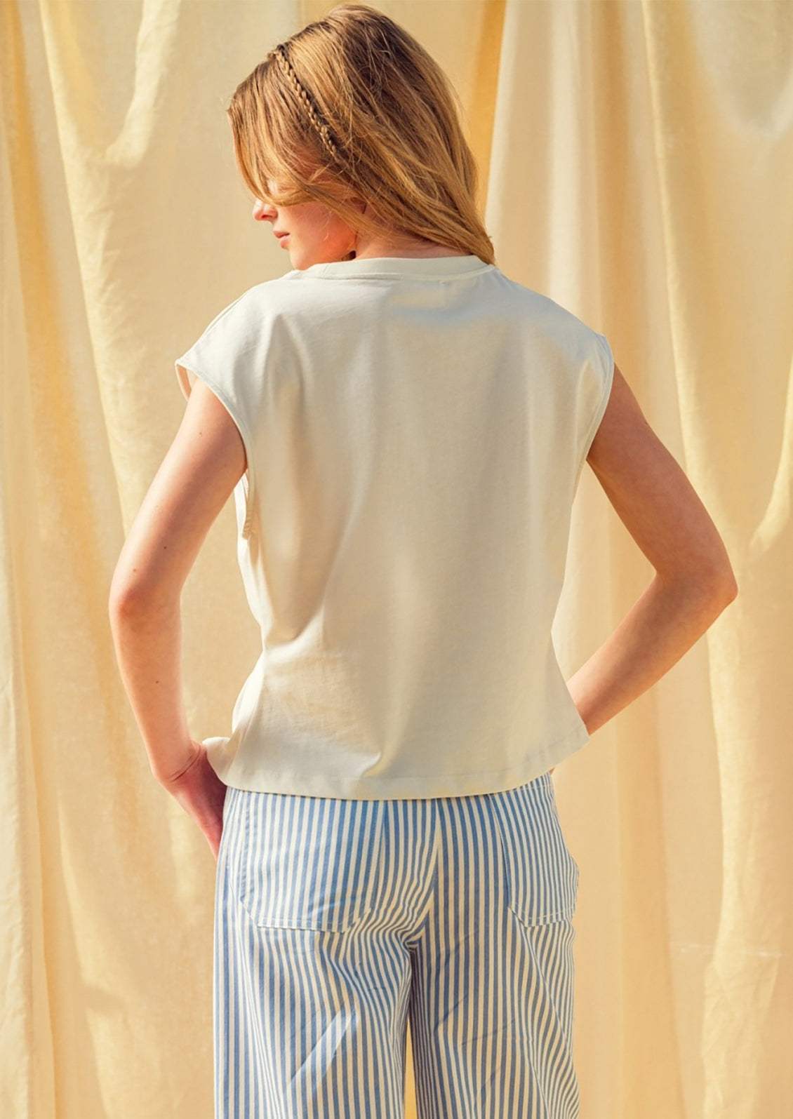 Person wearing a white sleeveless top and striped pants against a beige curtain background