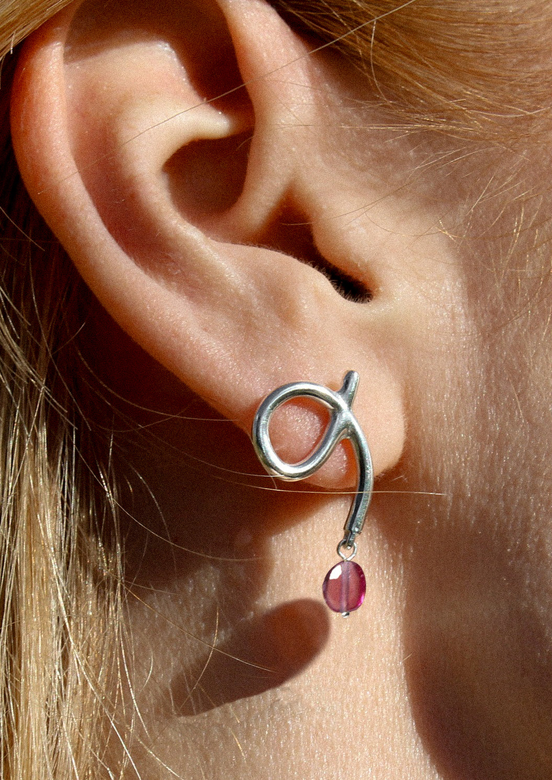 A person wearing a pair of silver loop shaped earrings with dangling garnet bead.