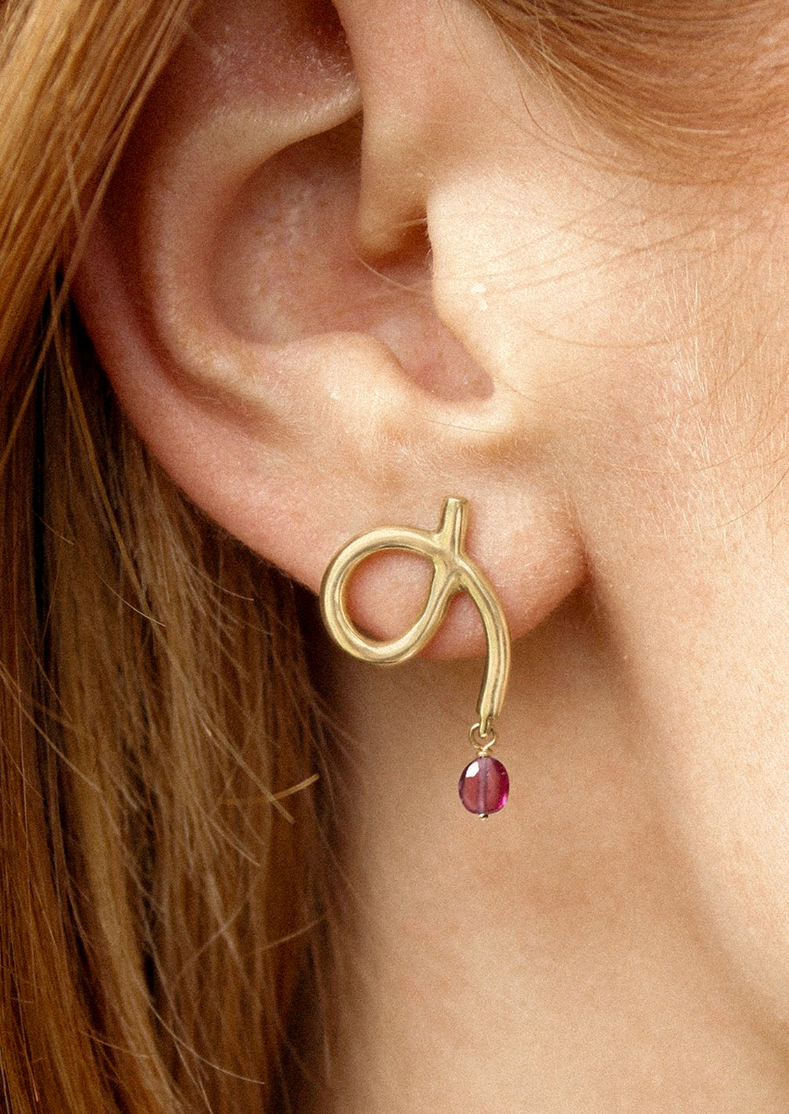 A person wearing a pair of gold loop shaped earrings with dangling garnet bead.