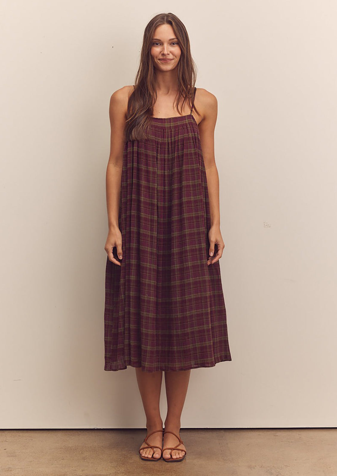 A person wearing a midi length dress in burgundy and green plaid pattern with thin straps.