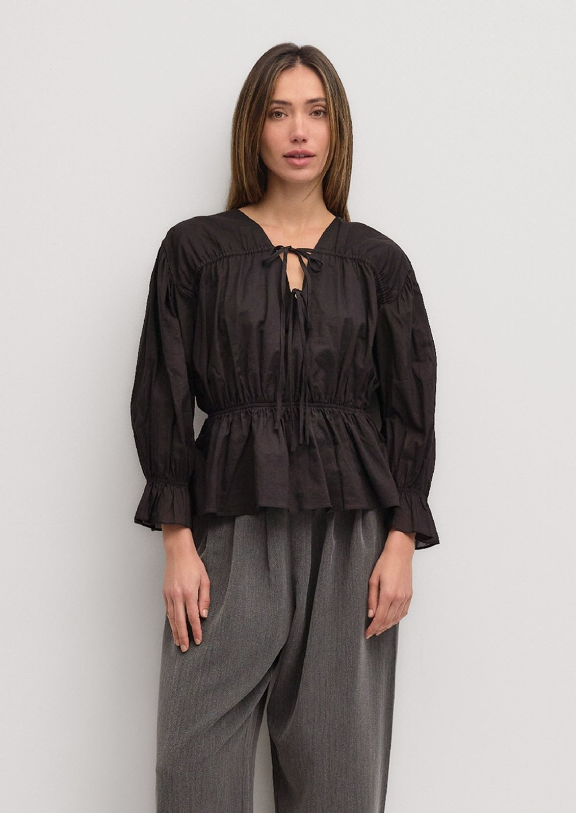 A woman wearing a black cotton blouse with ruching and bow tie detail at front.