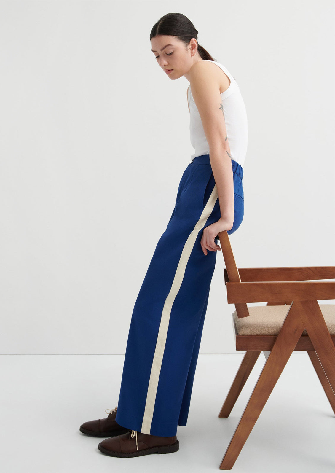 Woman in blue pants with a white stripe standing next to a wooden chair on a white background