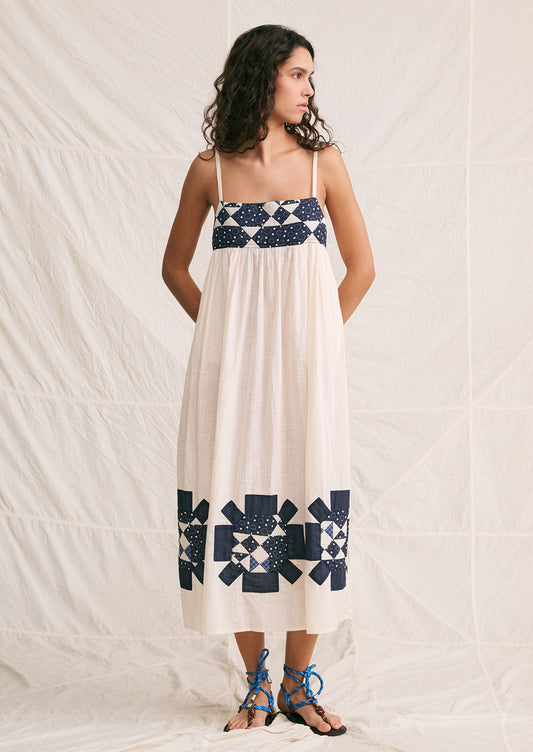 A woman wearing a long white sundress with indigo patchwork quilting details.