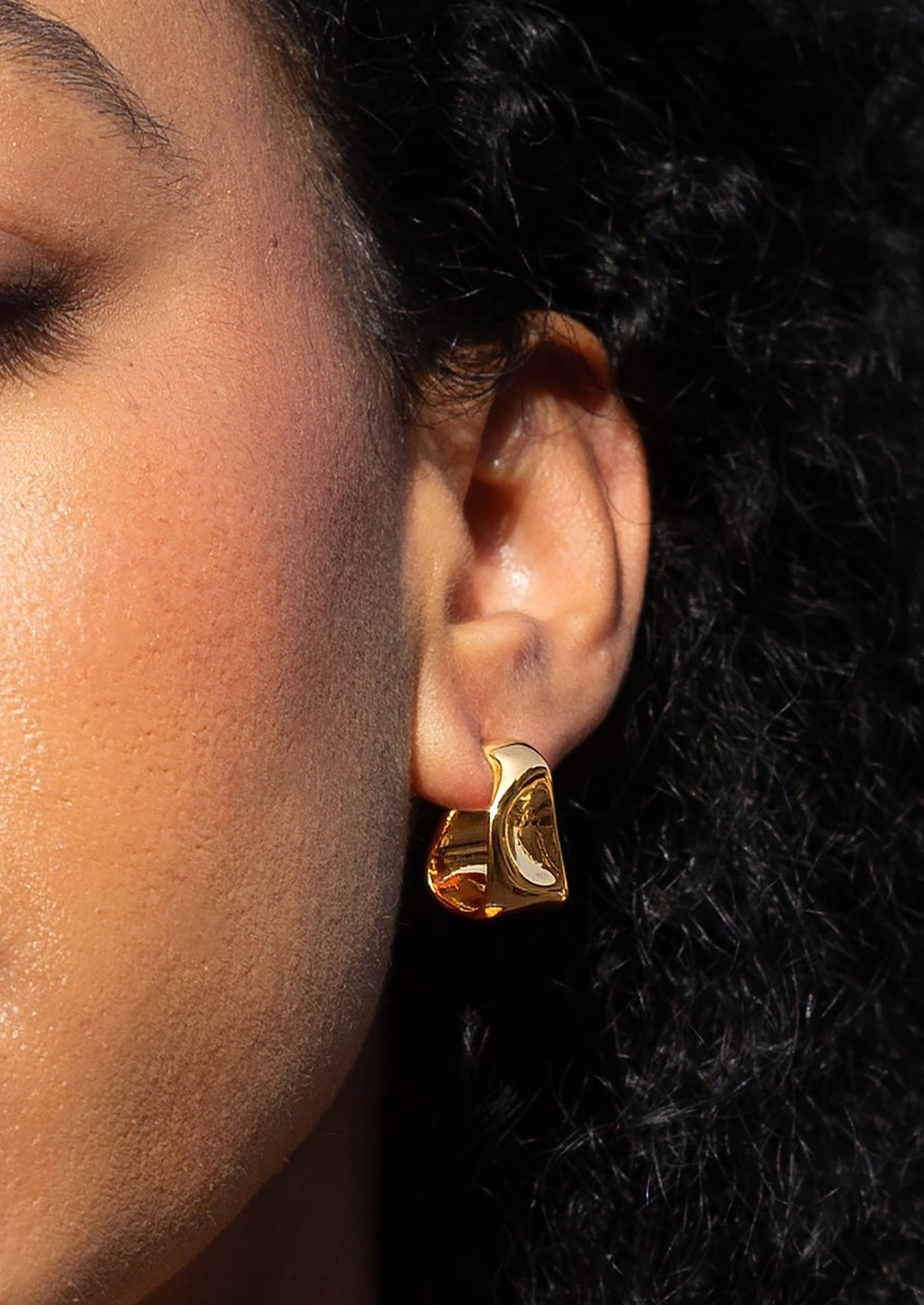 A person wearing a pair of asymmetrical gold hoop earrings with hammered wavy texture.