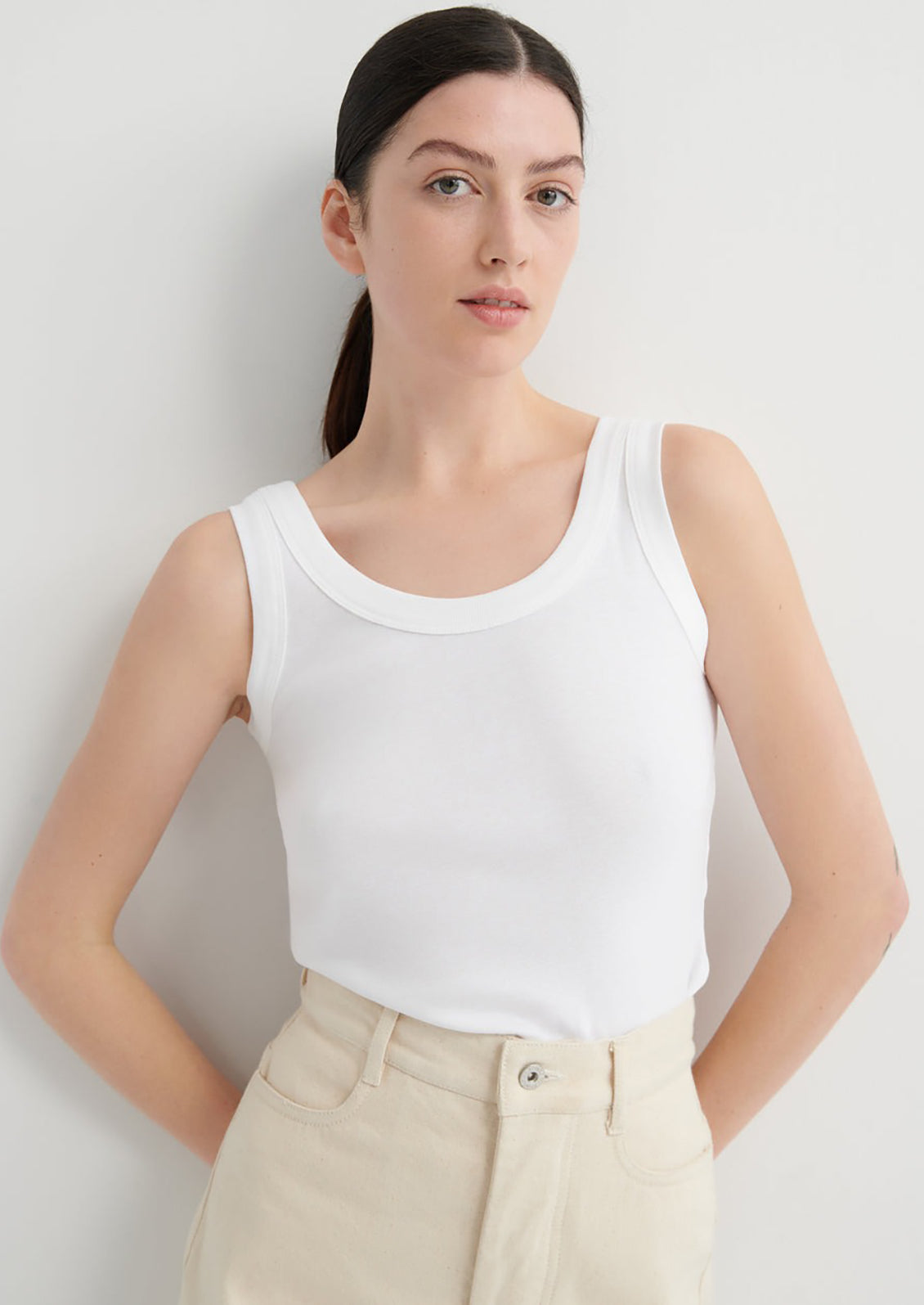 Woman wearing a white tank top and beige pants against a light gray background