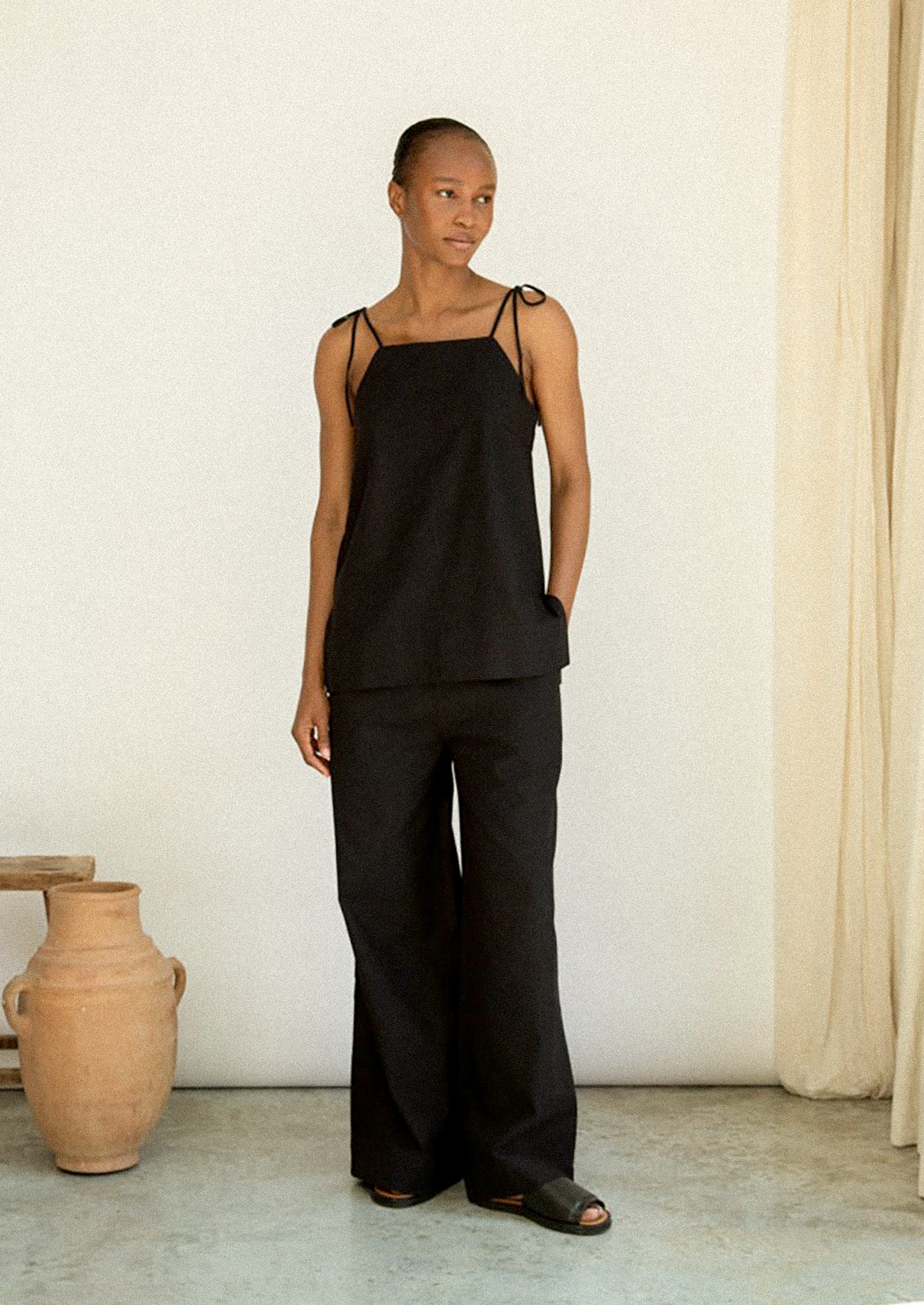 Woman wearing a black sleeveless top and wide-leg pants in a minimalistic setting.
