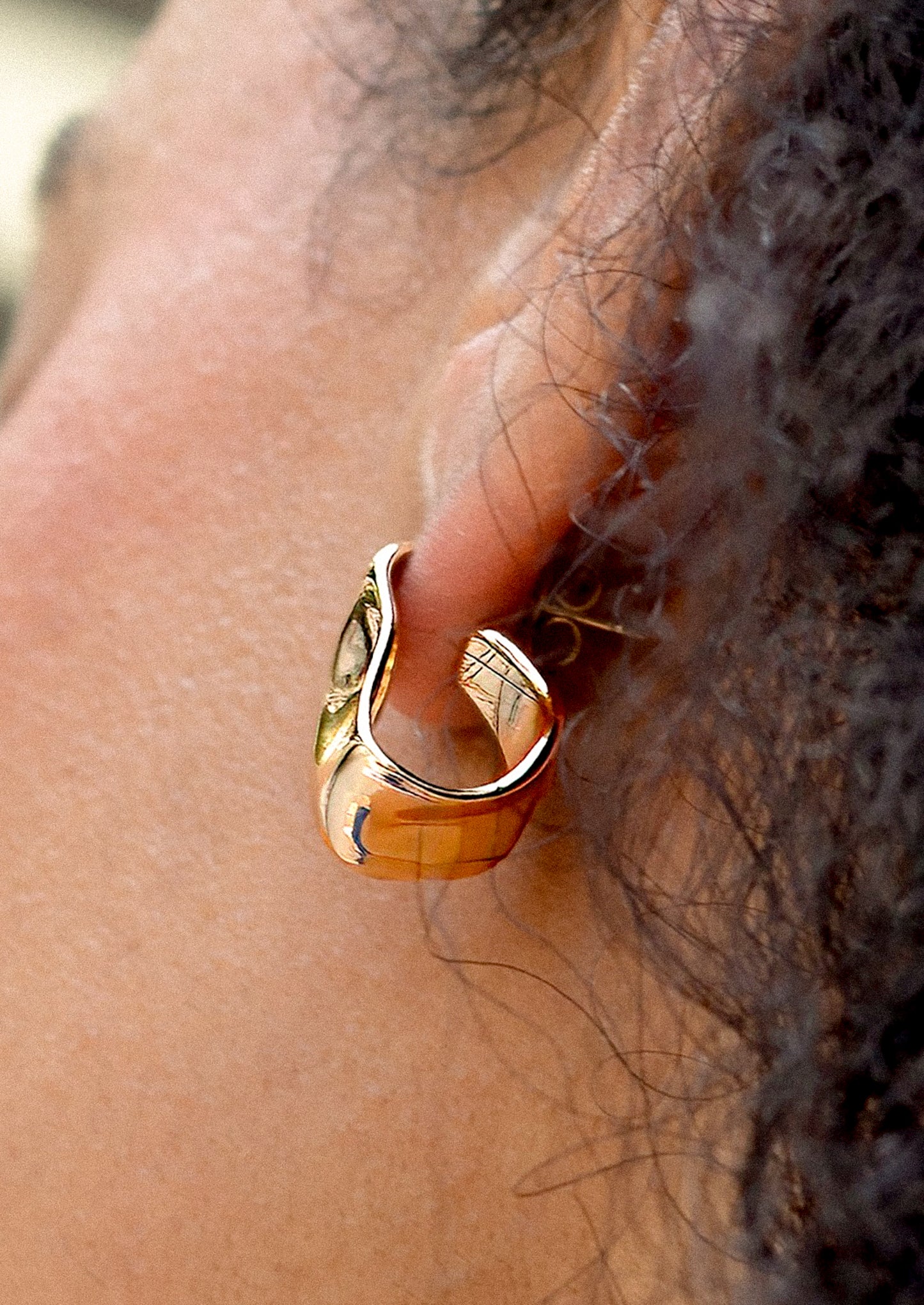 A person wearing a pair of asymmetrical gold hoop earrings with hammered wavy texture.
