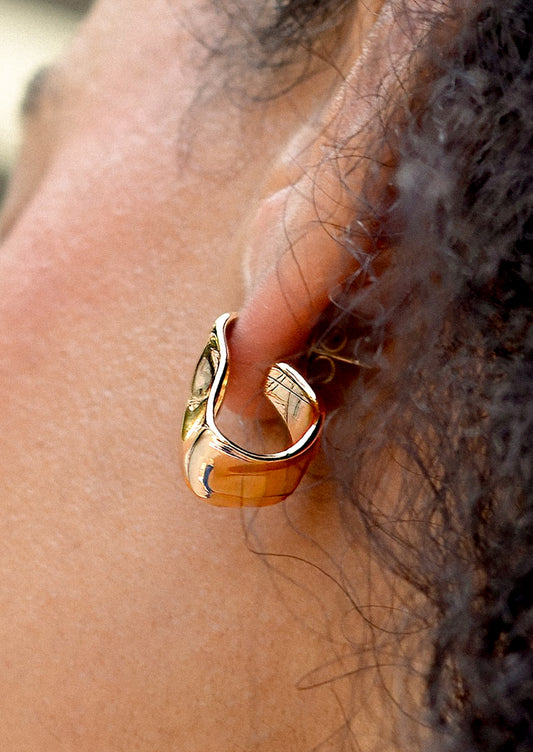 A person wearing a pair of asymmetrical gold hoop earrings with hammered wavy texture.