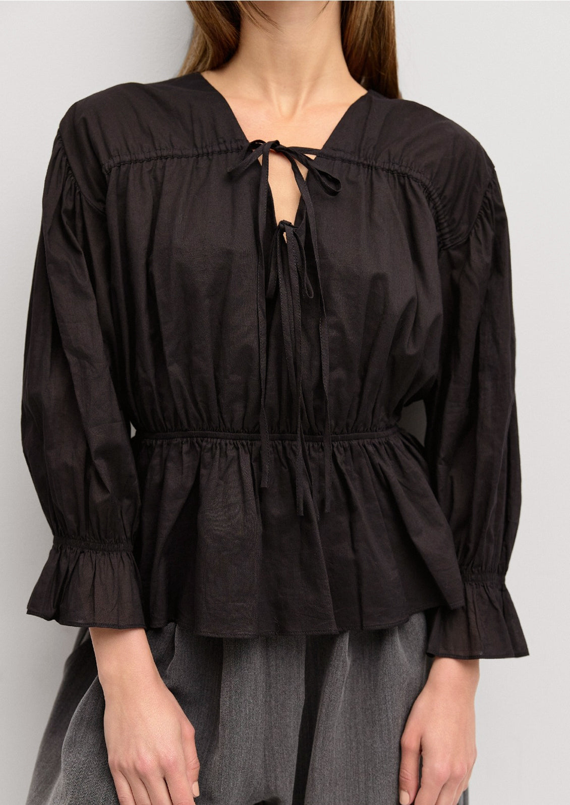 A woman wearing a black cotton blouse with ruching and bow tie detail at front.