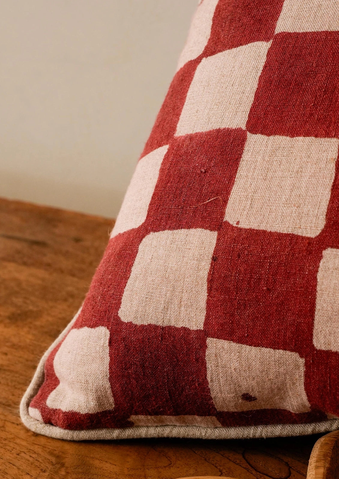 A close-up of a square throw pillow with a red and beige checkerboard pattern, resting on a rustic wooden surface.