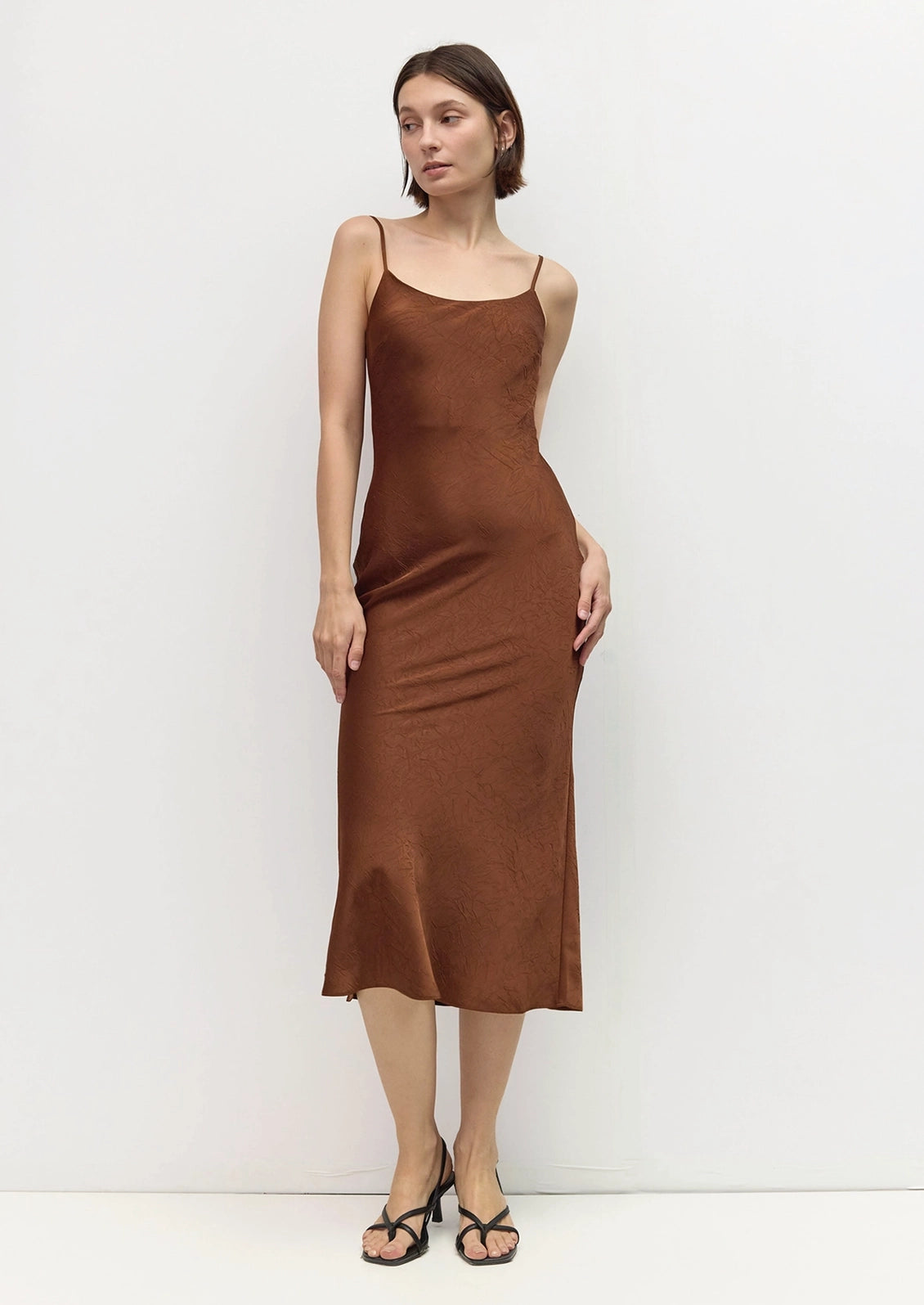 A woman modeling a brown satin midi slip dress with spaghetti straps, standing against a plain white studio background.