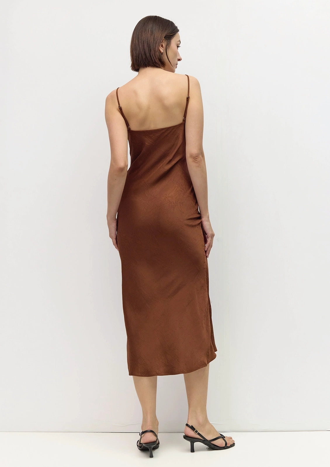 A woman viewed from the back wearing a brown satin slip dress with thin straps and black strappy heels, standing against a plain white wall.