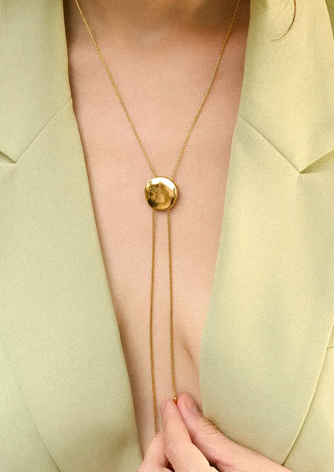 A close-up of a model wearing a gold lariat necklace with a round pendant and a long drop chain.