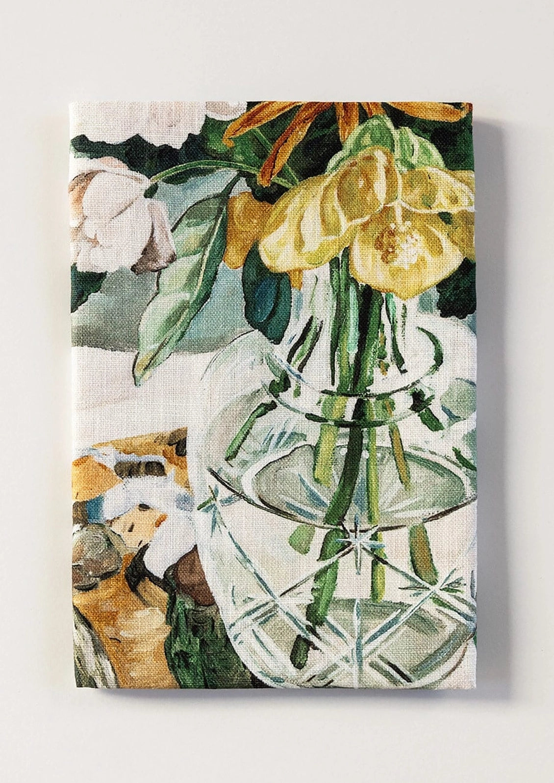 A tea towel featuring a printed still life painting of yellow flowers in a clear glass vase with green leaves on a light background.