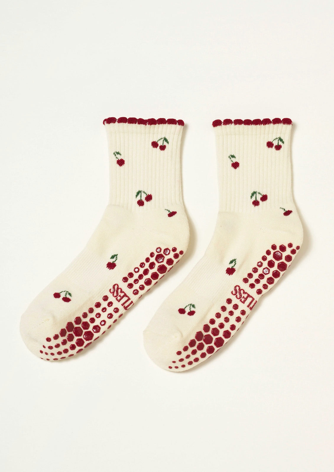 A pair of ribbed cream socks with red cherries embroidered on them, a ruffled trim, and grips at the bottom to prevent slipping.