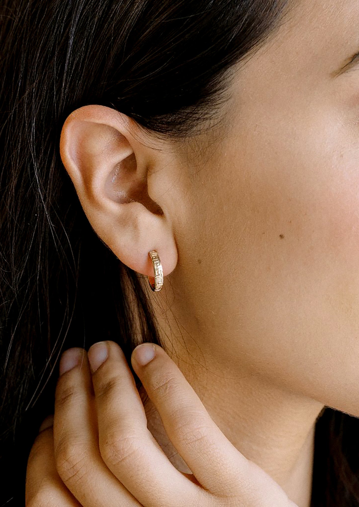 A close-up of a model's ear wearing a small gold hoop earrings.
