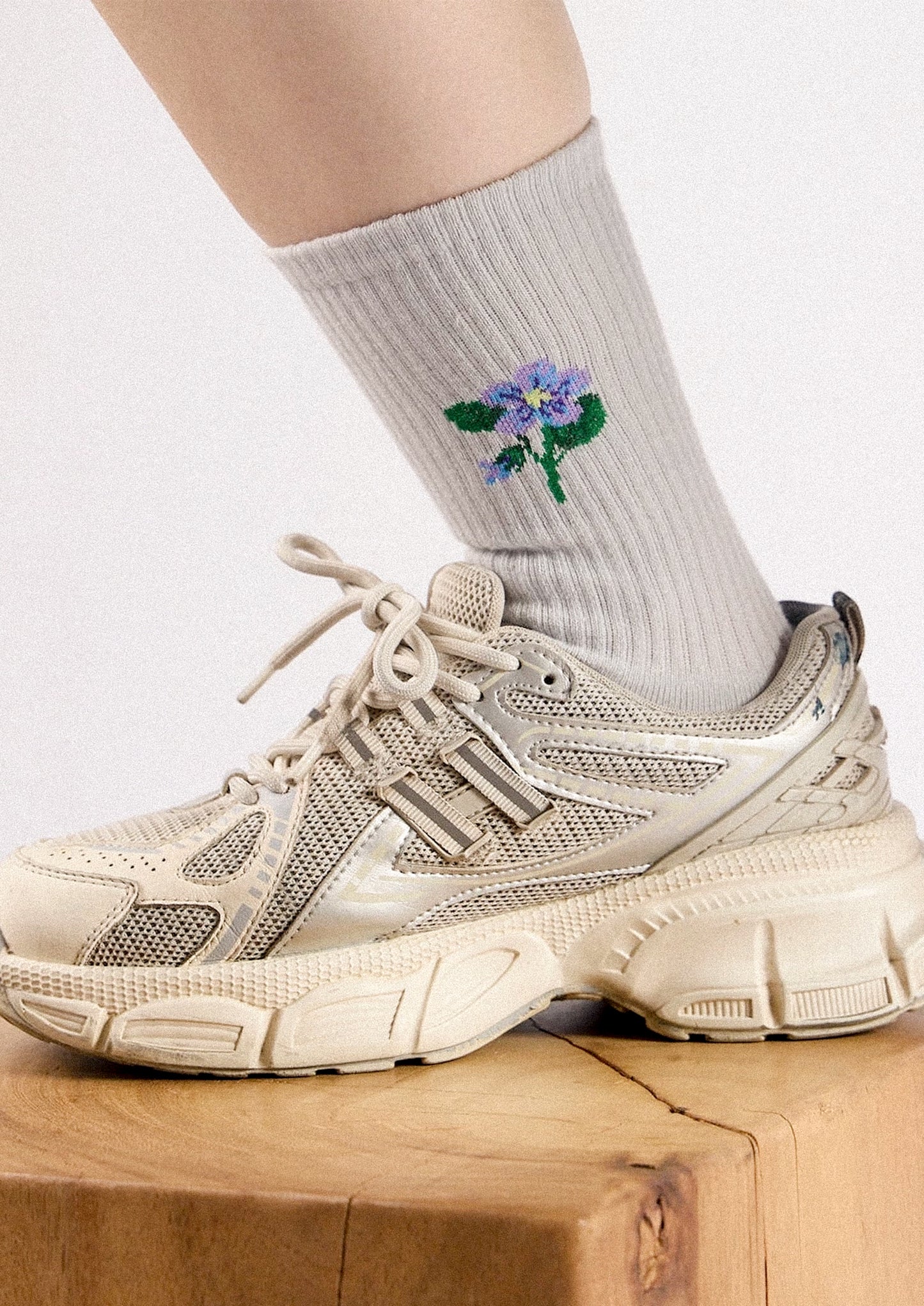 A close-up of a person's lower leg wearing a ribbed light gray sock with a small embroidered purple flower and green leaves, paired with a chunky beige sneaker on a wooden block against a light background.