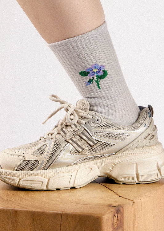 A close-up of a person's lower leg wearing a ribbed light gray sock with a small embroidered purple flower and green leaves, paired with a chunky beige sneaker on a wooden block against a light background.