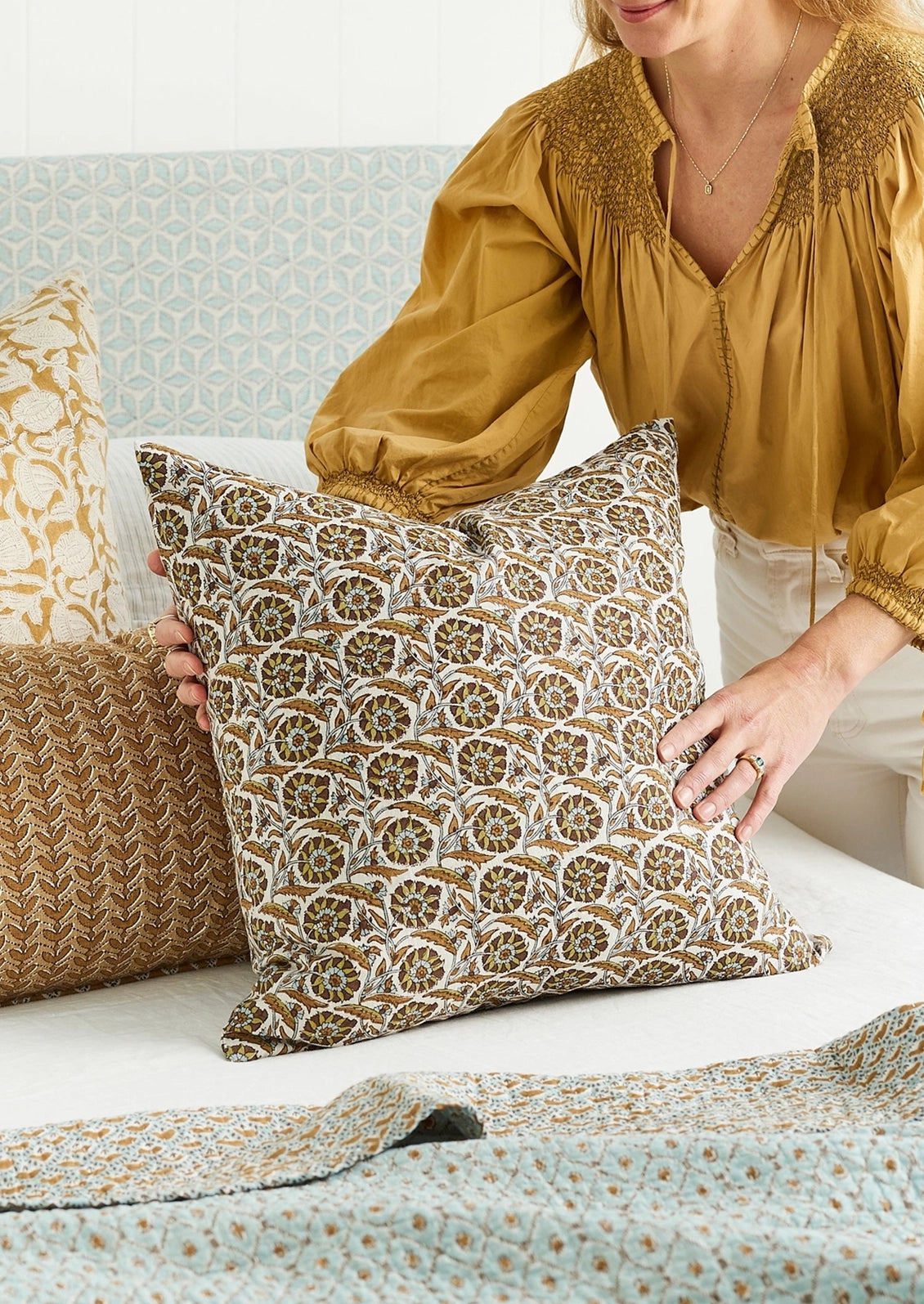 A woman in a mustard yellow blouse holds a square throw pillow with an intricate brown, gold, and white circular medallion pattern, styled on a bed with matching patterned textiles.