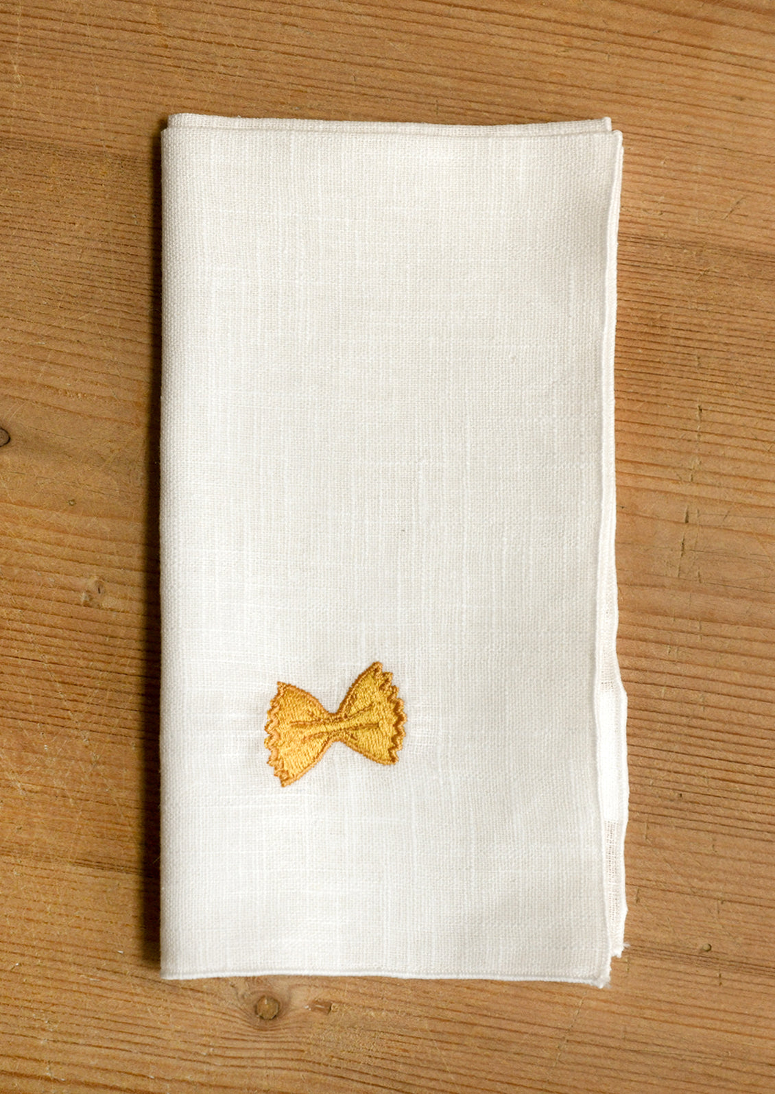 An off-white linen napkin folded flat on a wooden surface, featuring a small embroidered gold bow tie pasta near the bottom.