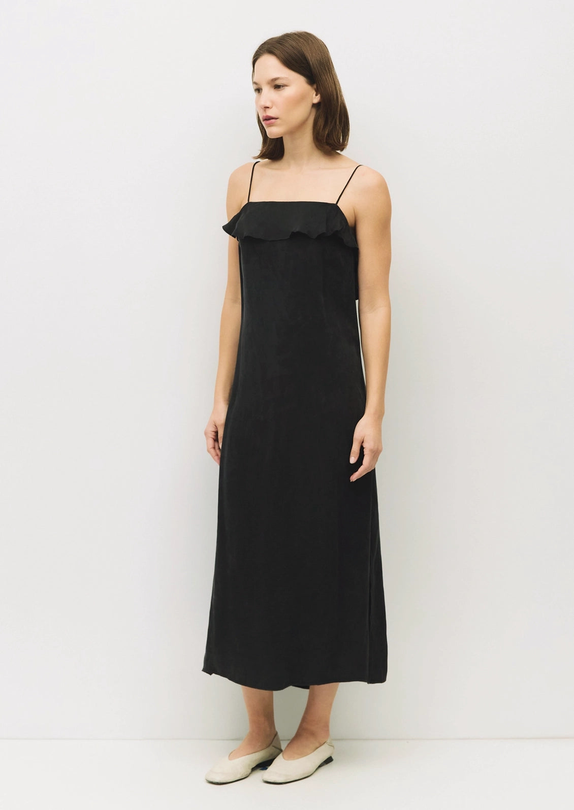 A woman modeling a black midi slip dress with thin spaghetti straps and a ruffled neckline, paired with light beige flat shoes, set against a plain white studio background.