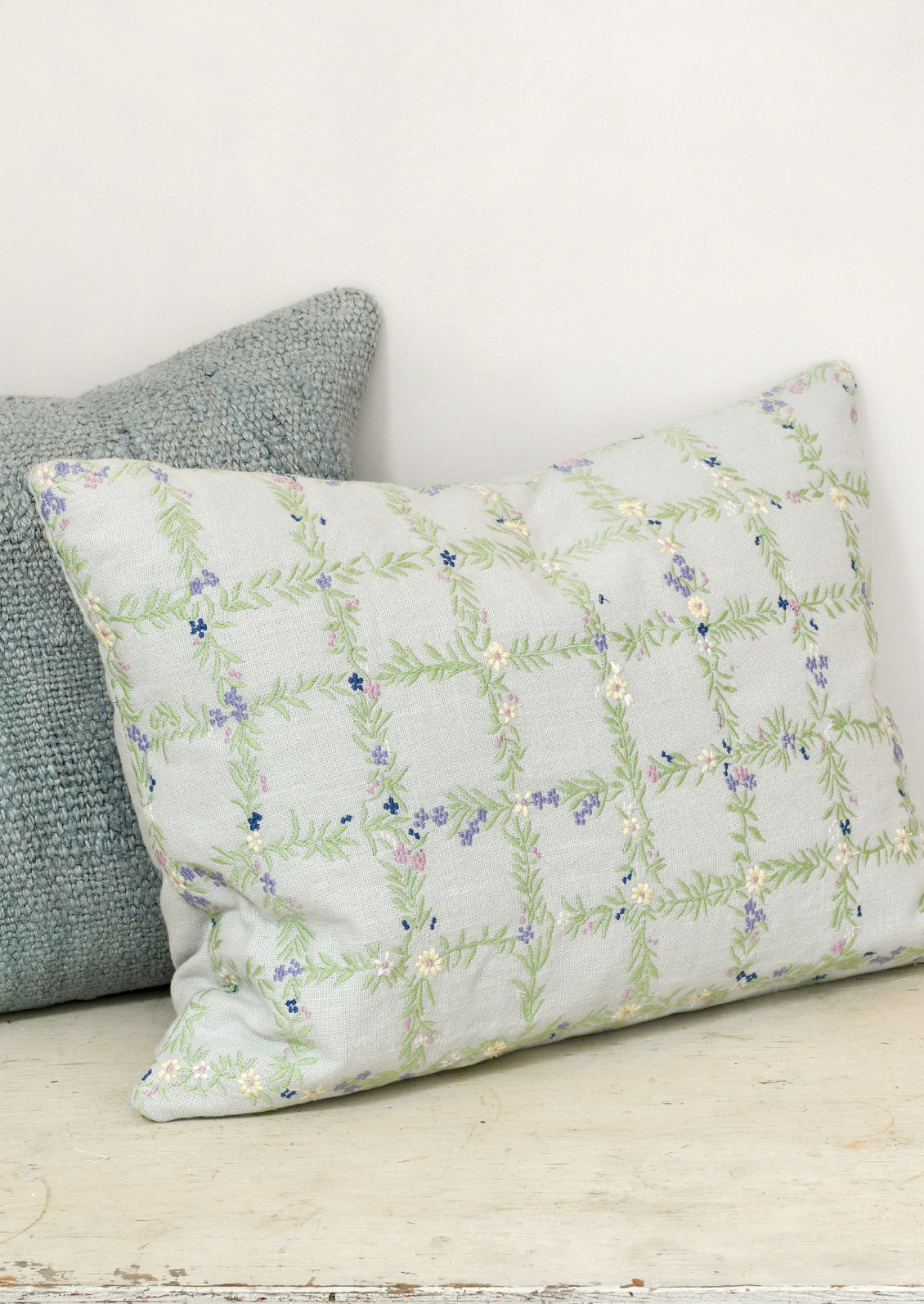 A light blue pillow with an embroidered floral grid pattern sitting on a table and leaning on a blue hemp pillow.