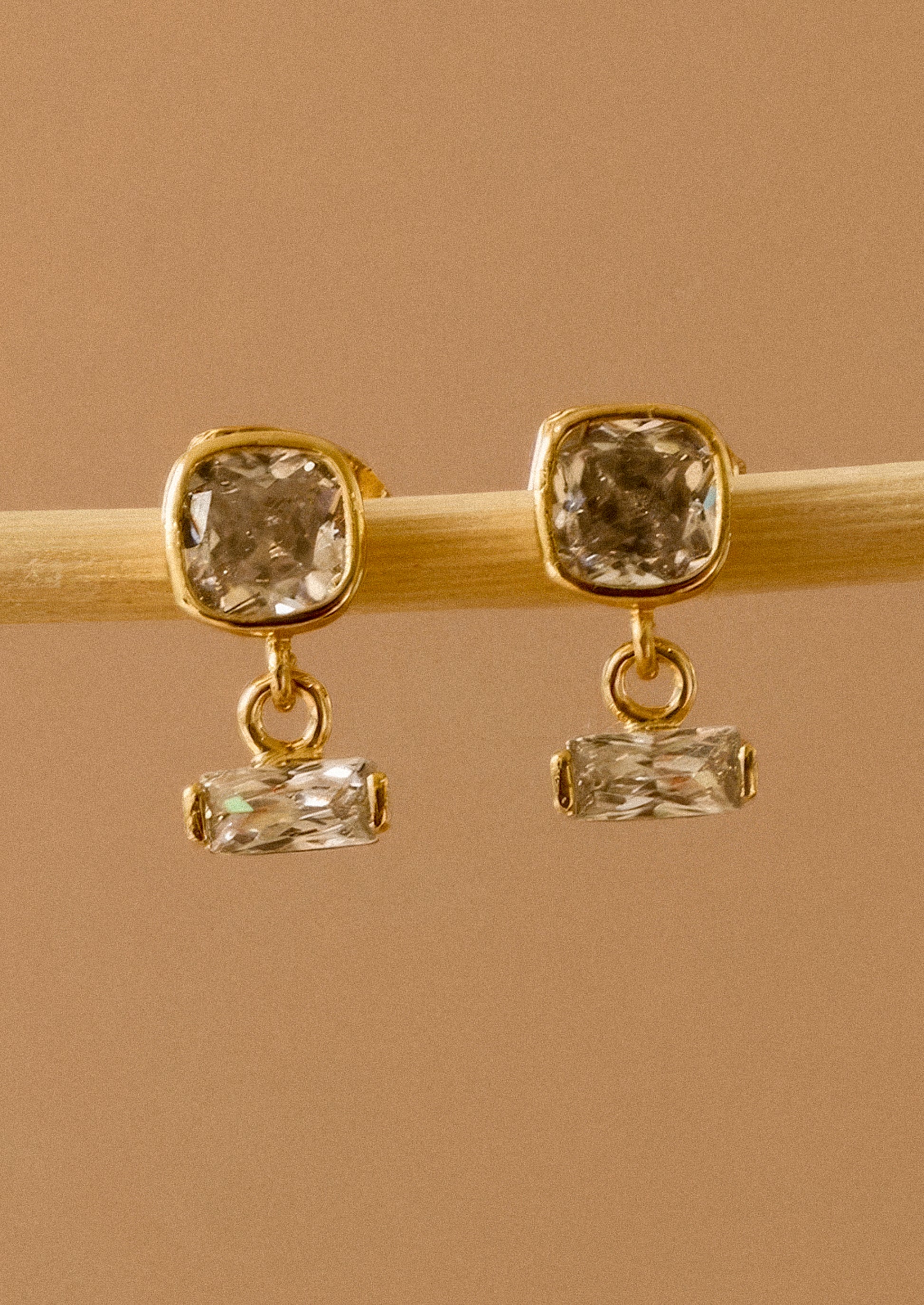 A pair of gold drop earrings with square cushion-cut clear crystals on the top and smaller rectangular clear crystals below, displayed on a light wooden stick against a warm tan background.