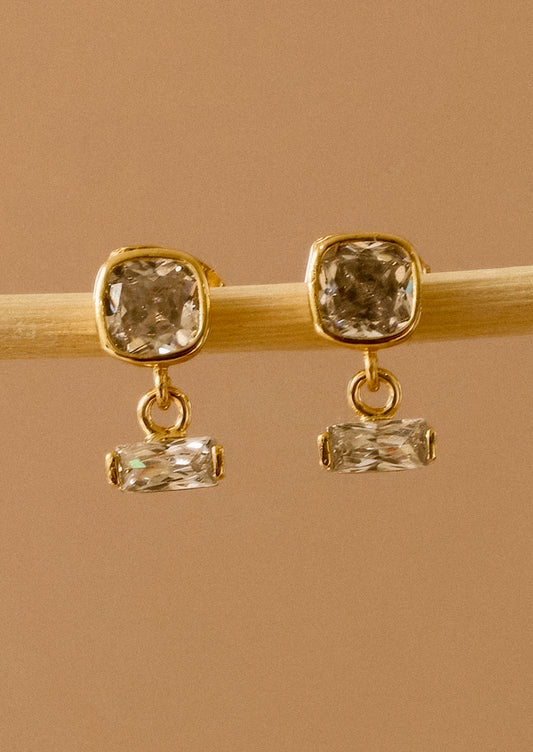 A pair of gold drop earrings with square cushion-cut clear crystals on the top and smaller rectangular clear crystals below, displayed on a light wooden stick against a warm tan background.