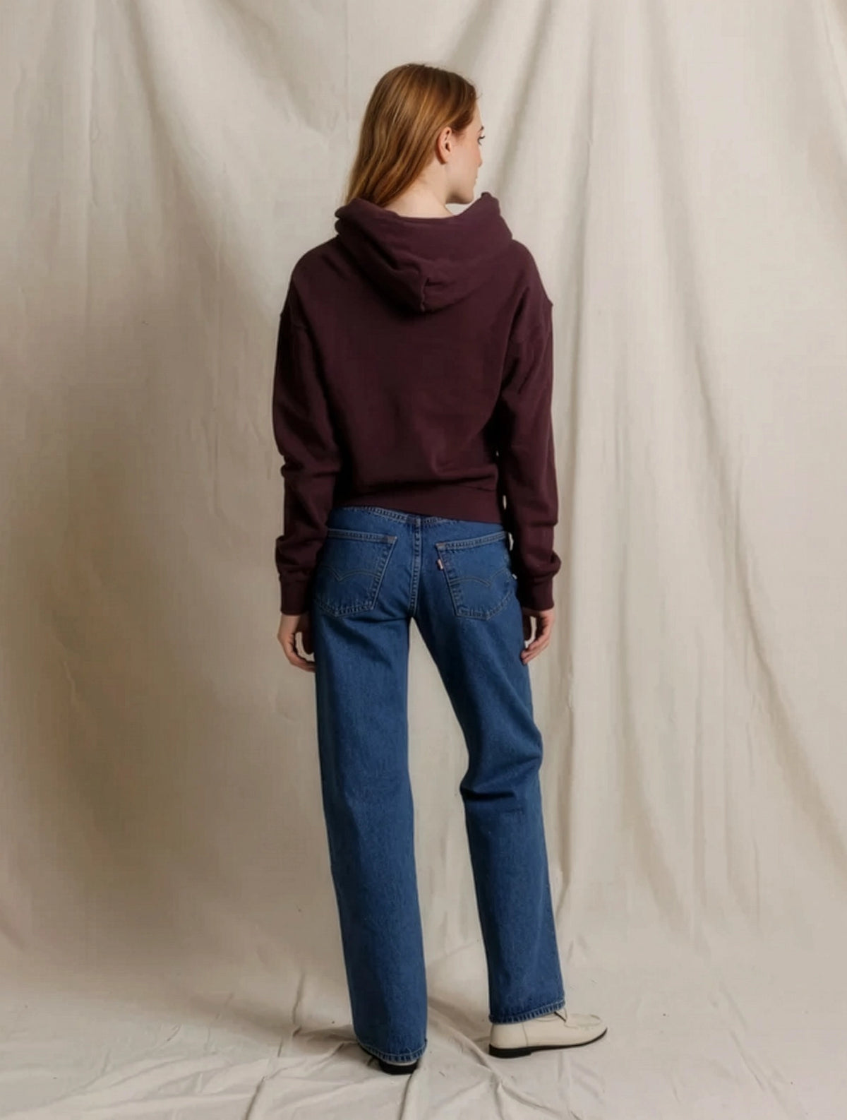 A woman is shown from the back wearing a dark plum-purple cropped hoodie and blue jeans, standing against a draped beige fabric backdrop.
