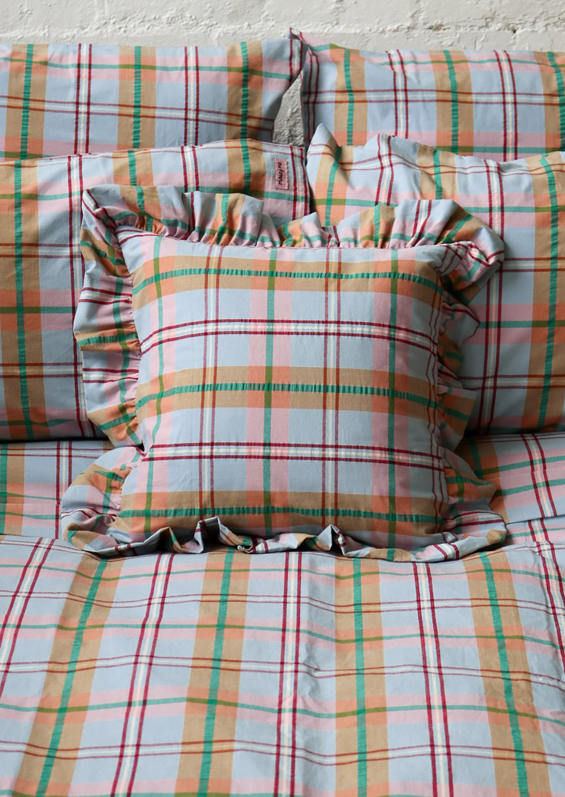 A bed styled with a pastel plaid bedding set and matching pillows, featuring a central ruffled pillow. The plaid pattern contains soft orange, green, red, and light blue lines on a white base.