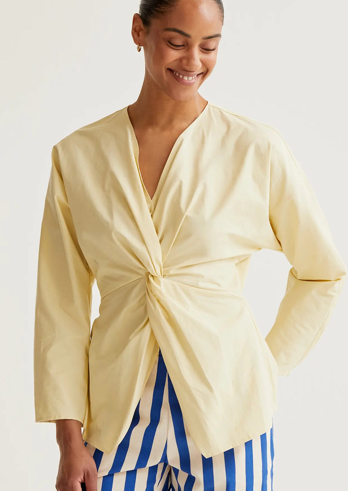Woman wearing a light yellow top with a knot detail and blue striped pants on a white background