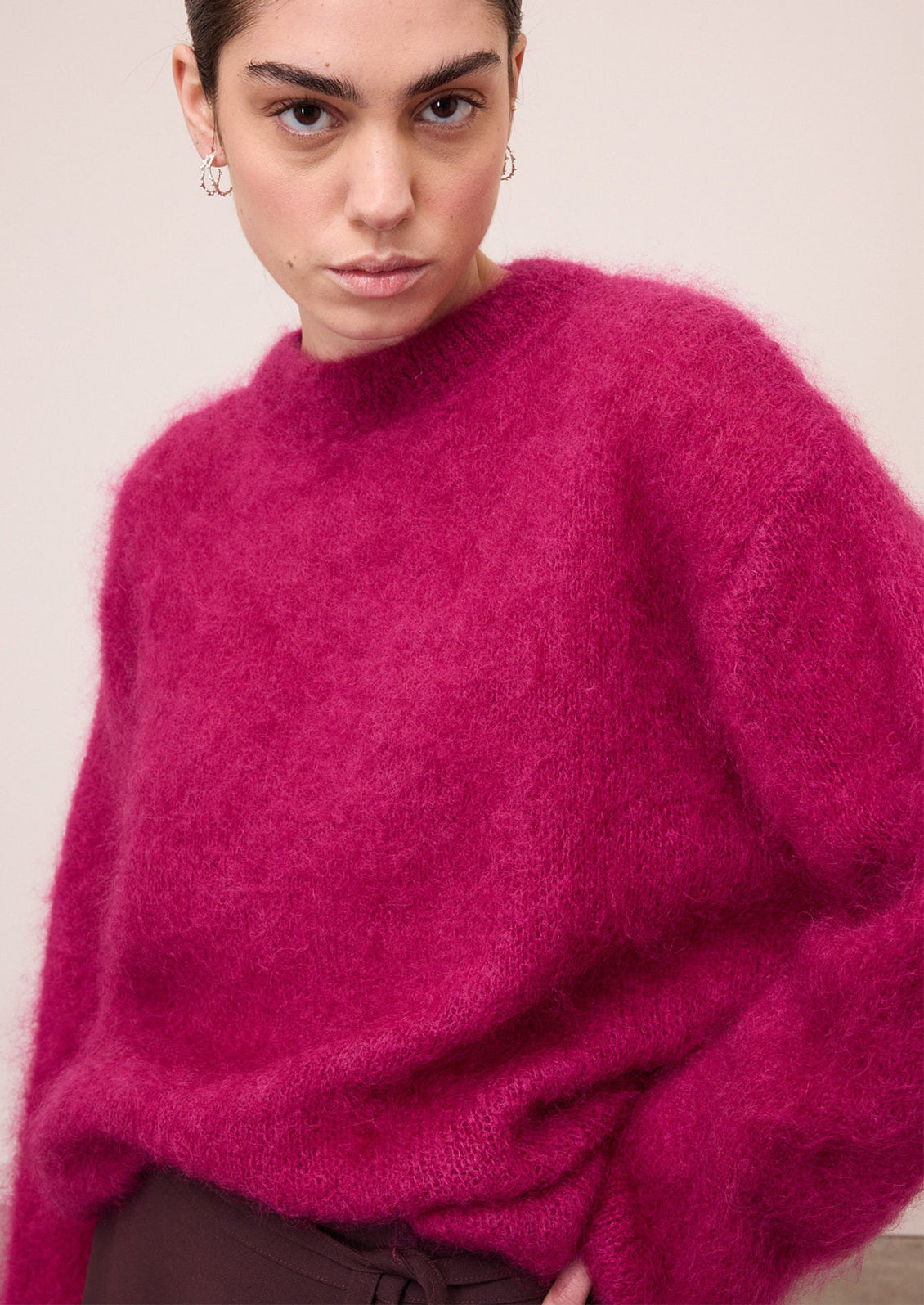 A close-up of a model wearing a fuzzy, mohair-like knit sweater in a vibrant magenta pink color, paired with dark brown pants against a plain beige background.