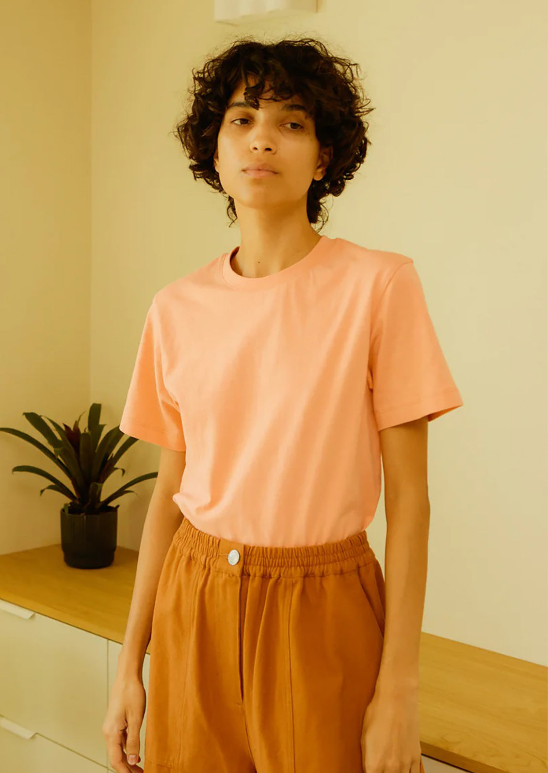 Person wearing a peach-colored t-shirt and brown pants standing indoors.