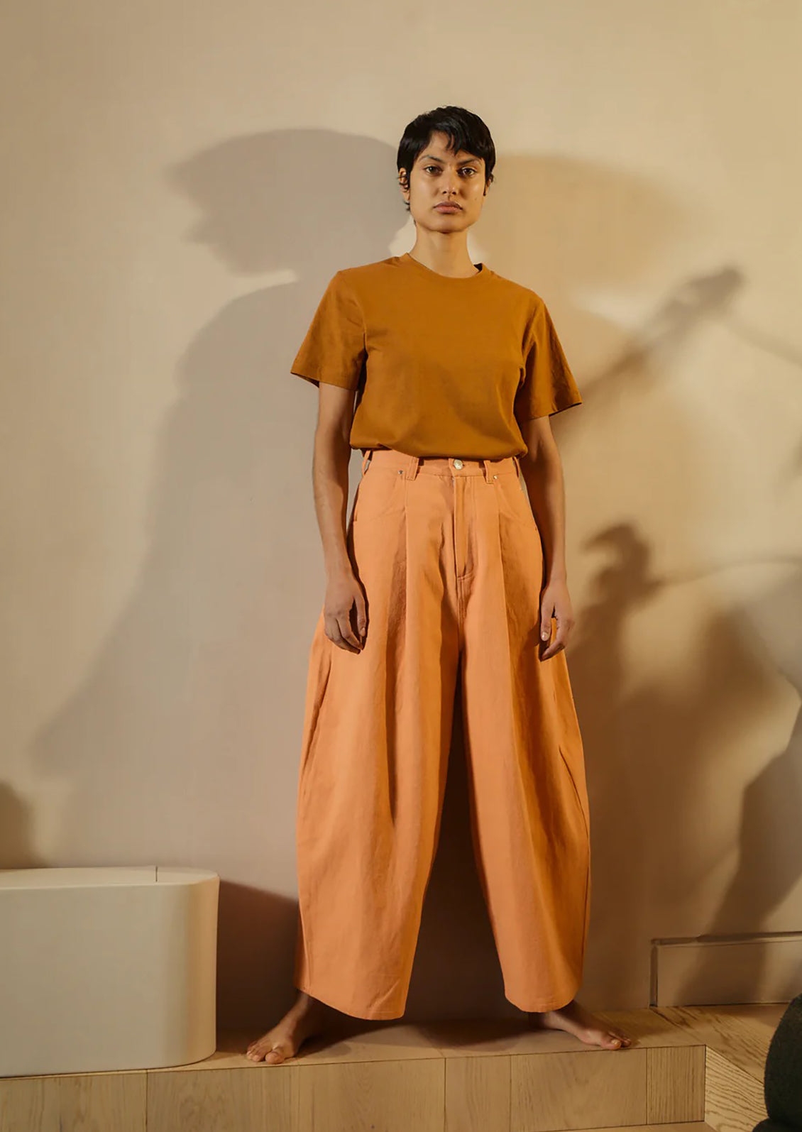 Person wearing a mustard yellow top and orange pants standing against a beige wall.