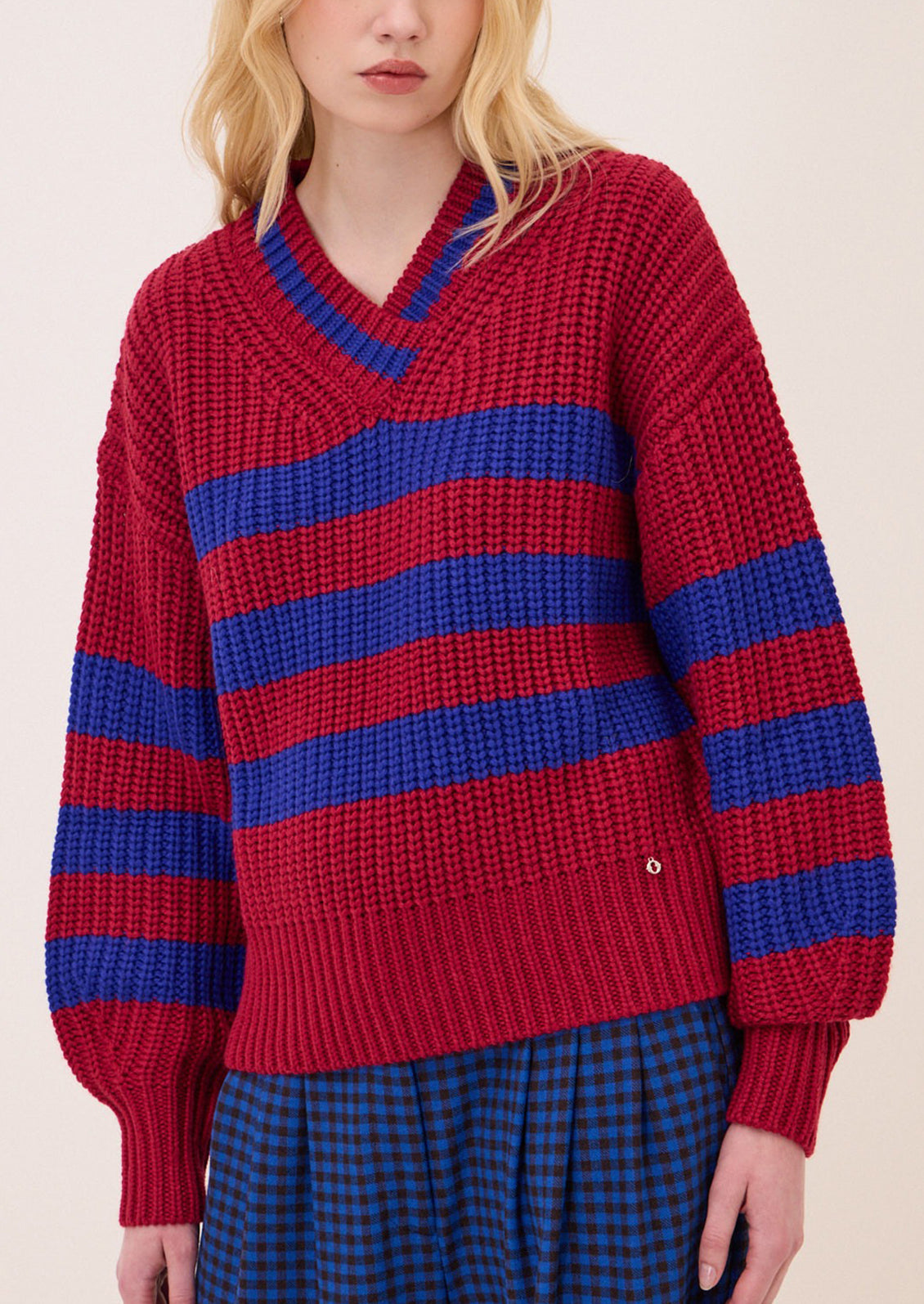 A model wearing a chunky knit V-neck sweater with horizontal red and blue stripes, paired with a blue and black checkered skirt, against a plain light beige background.