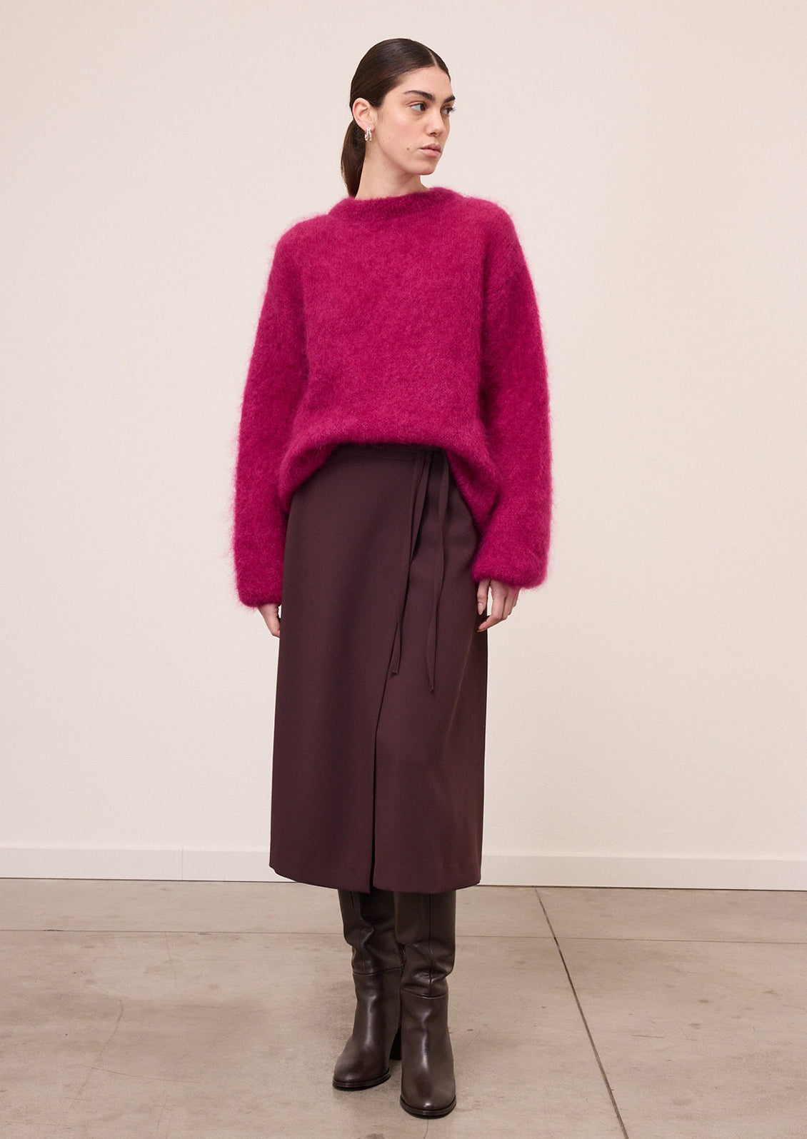 A model wearing a fuzzy, mohair-like knit sweater in a vibrant magenta pink, paired with dark brown trousers, against a plain off-white wall.