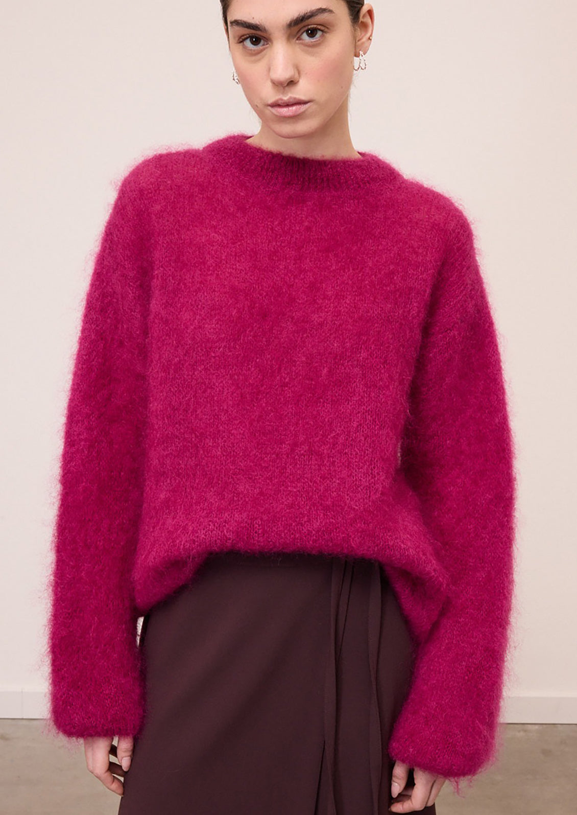 A model wearing a fuzzy, mohair-like knit sweater in a vibrant magenta pink, paired with dark brown trousers, against a plain off-white wall.