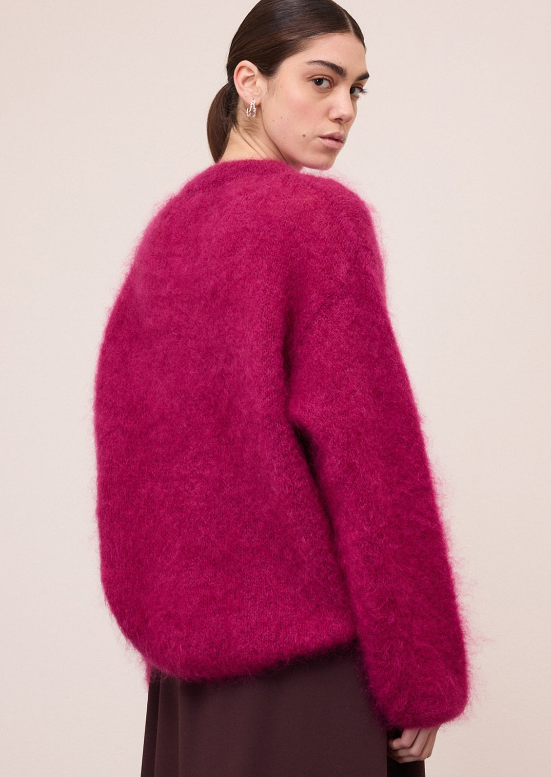 A model wearing a fuzzy, mohair-like knit sweater in a vibrant magenta pink, paired with dark brown trousers, against a plain off-white wall.