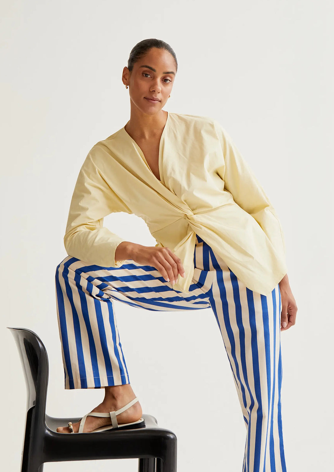 Woman wearing a yellow top and blue striped pants sitting on a black chair against a white background