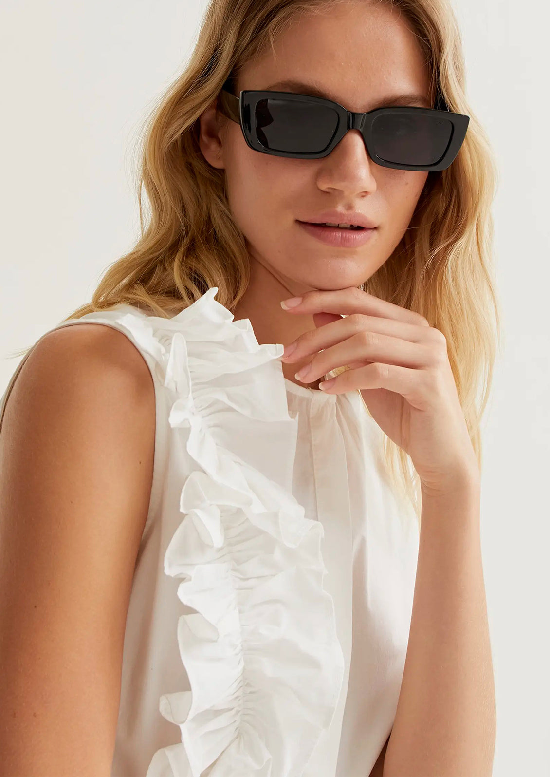 Woman wearing a white sleeveless top with ruffles and black sunglasses against a white background