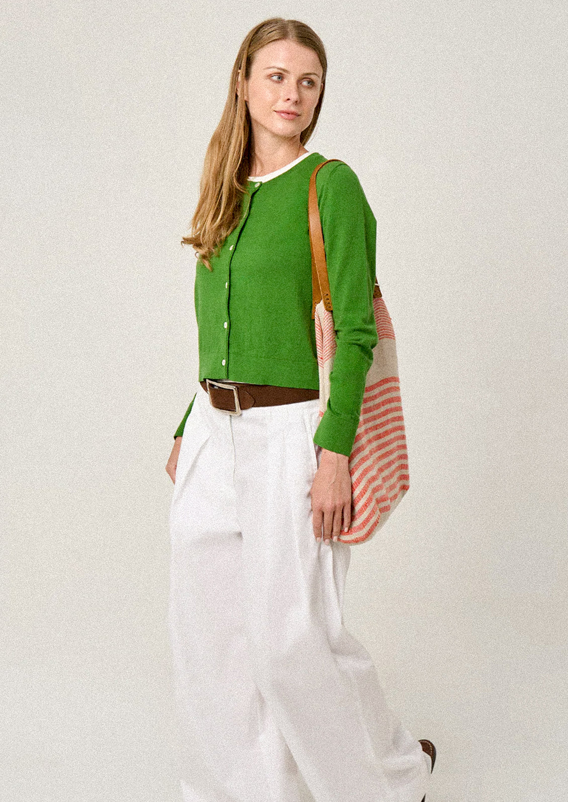 Woman wearing a green cardigan and white pants, holding a striped bag against a plain background