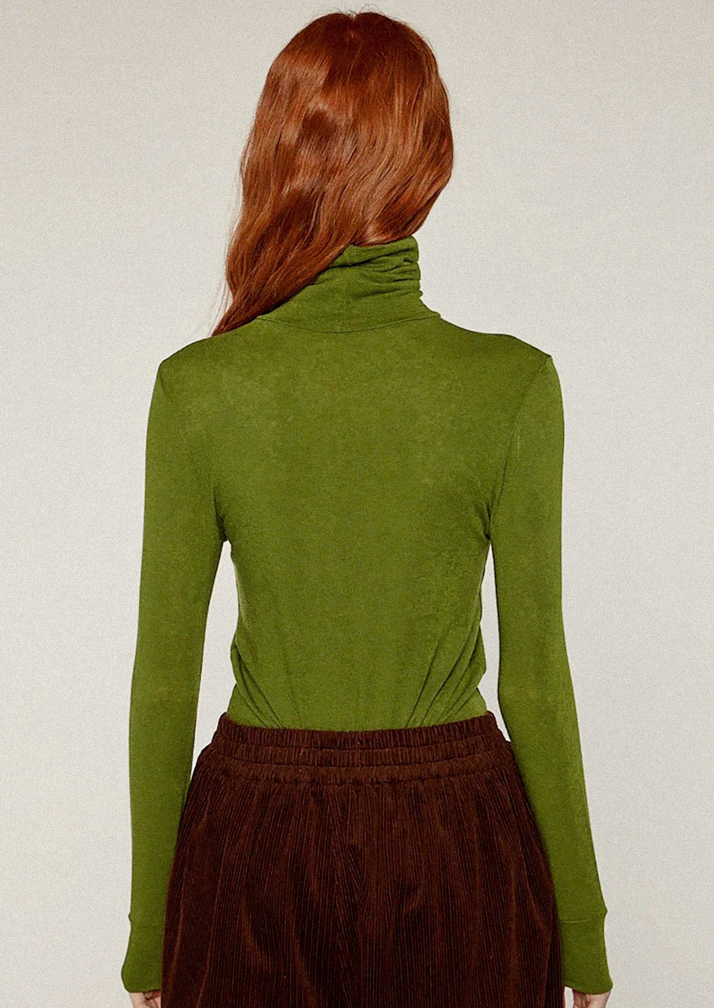 Person wearing a green turtleneck sweater and brown pants against a white background