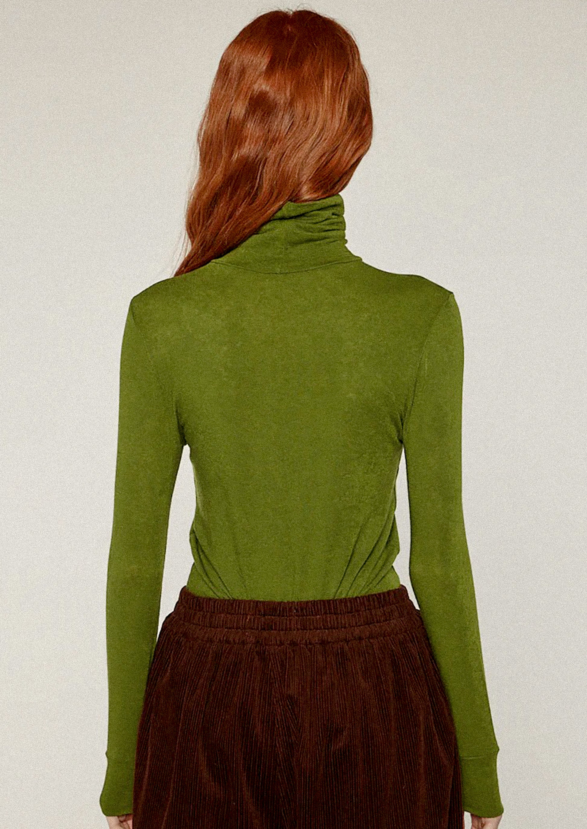 Person wearing a green turtleneck sweater and brown pants against a white background