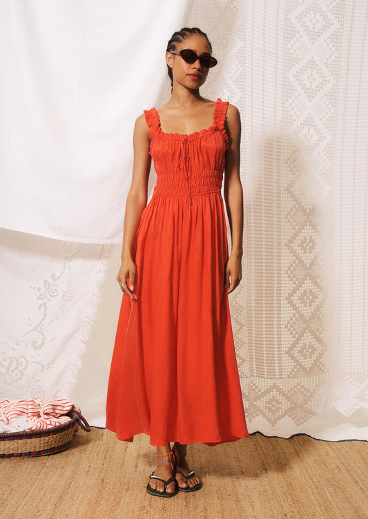 A woman modeling a bright orange-red midi dress with ruffled straps, a gathered bodice, and a flowing skirt.