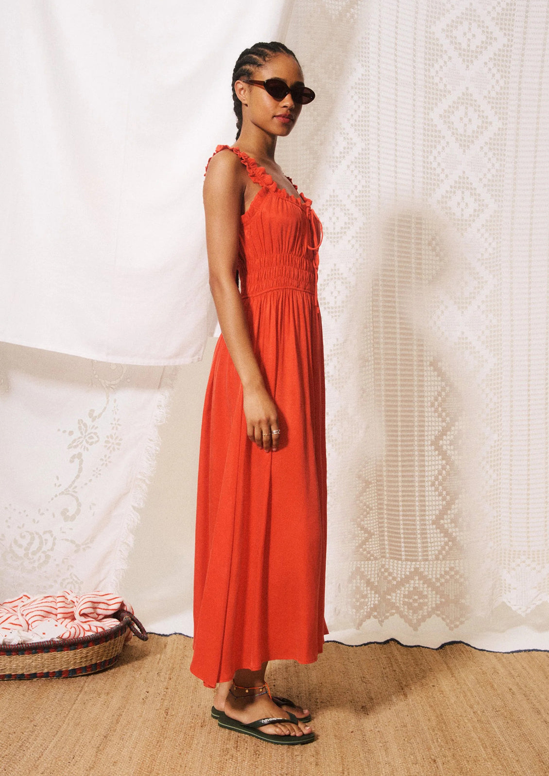 A woman modeling a bright orange-red midi dress with ruffled straps, a gathered bodice, and a flowing skirt.