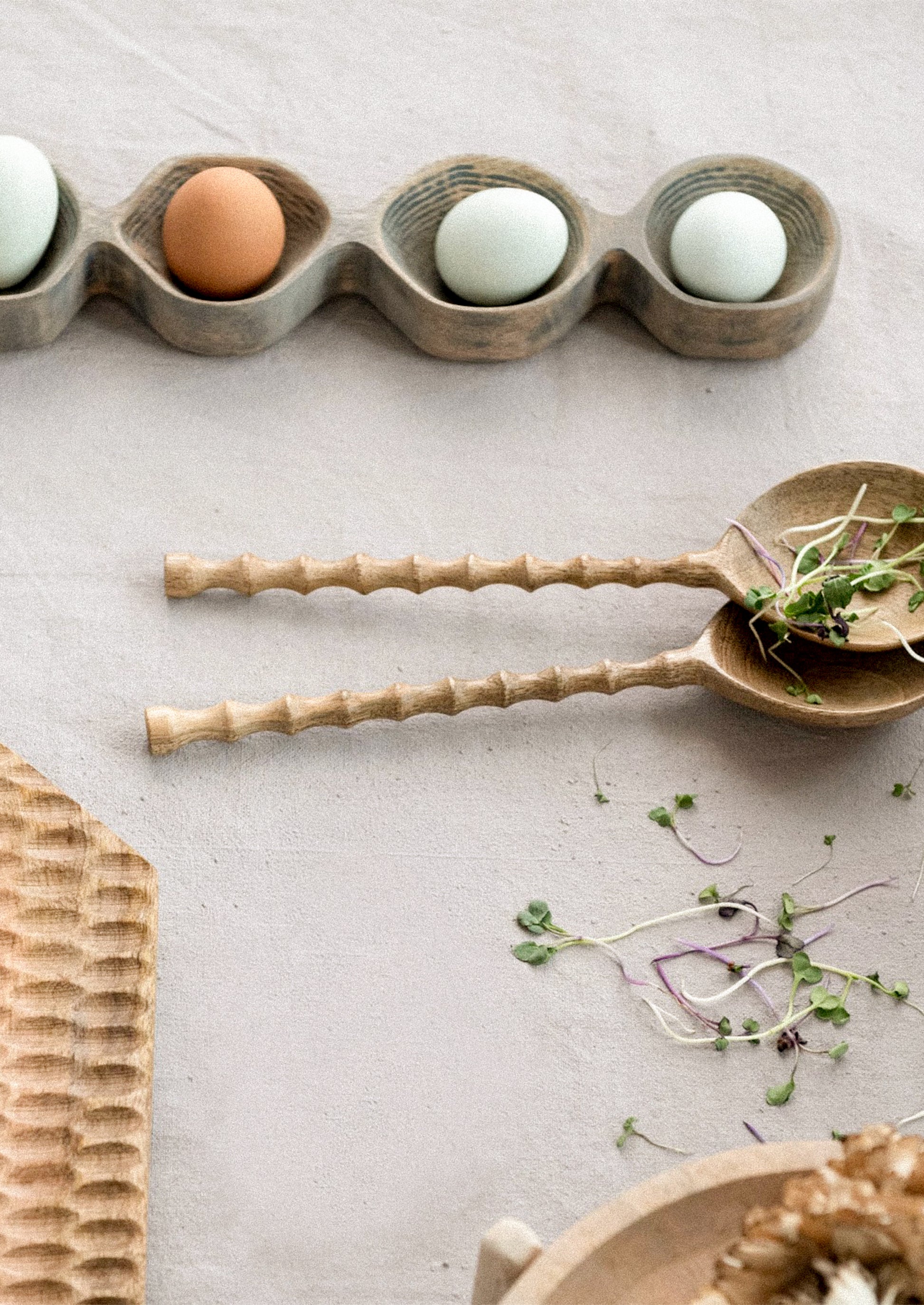 Wooden egg holder with eggs on a light surface, accompanied by a wooden spoon with sprouts.