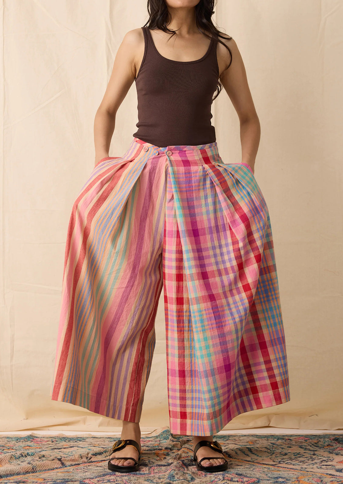 A model wearing wide-leg pleated trousers with a split design: one leg has vertical pink, orange, and yellow stripes, and the other has a pink, red, and blue plaid pattern. 
