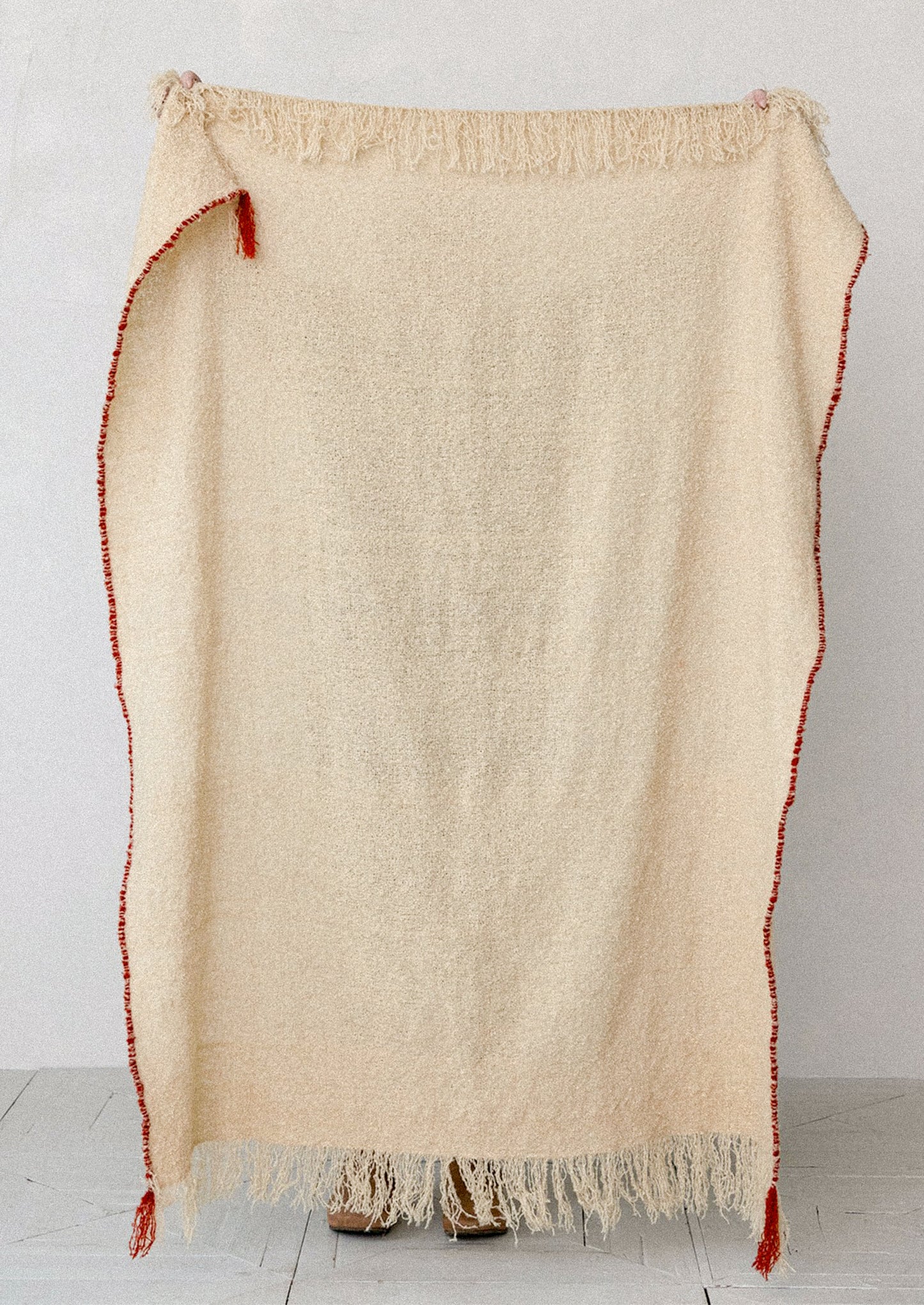 A cream-colored, textured woven blanket with fringe trim and red edging, displayed against a light gray wall.