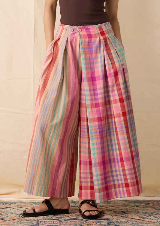 A person wearing wide-leg cropped pants with a split design: one leg has vertical pastel stripes in pink, blue, yellow, and green, and the other leg has a pink, red, and blue plaid pattern.
