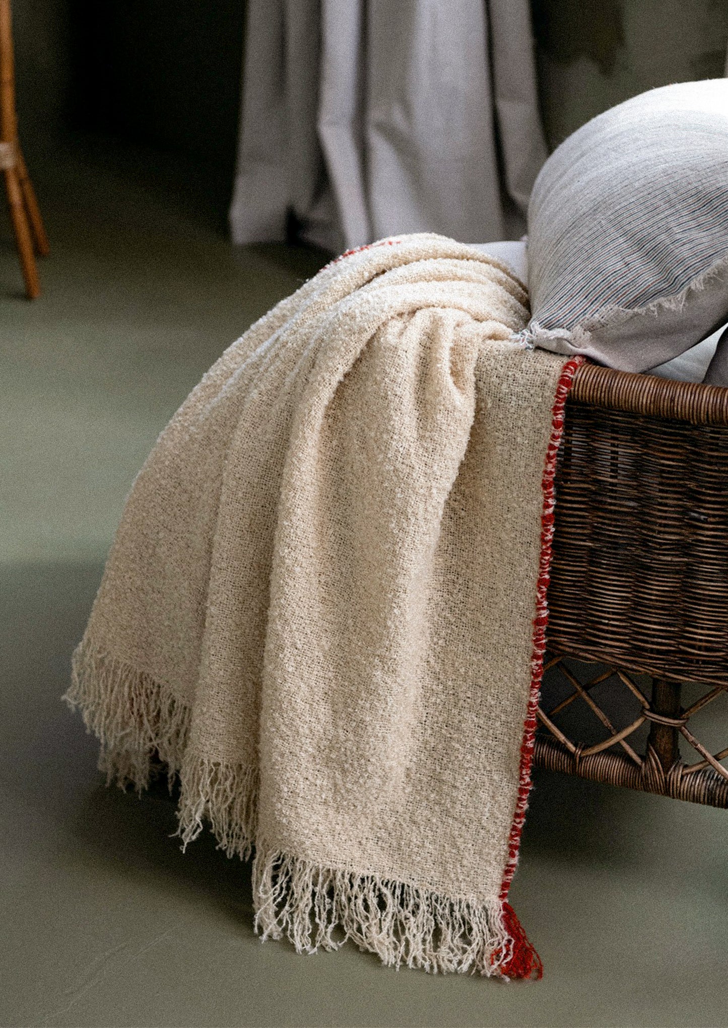 A cream-colored textured throw blanket with fringe and a red patterned edge, draped over a wicker chair next to a white pillow in a softly lit room.